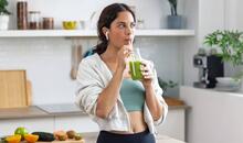 Mujer apoyada en la encimera de una cocina bebiendo un batido verde de frutas y verduras vestida de deporte, un ejemplo de microhábitos de alimentación saludable