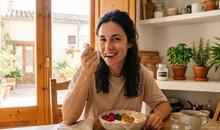 Una mujer sentada en la mesa de una cocina comiendo un bol de yogur natural, alimento rico en postbióticos, junto a frutas y semillas.