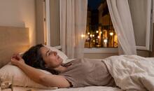 Mujer despierta tumbada en la cama junto a una ventana abierta durante una noche de verano, una escena habitual al buscar cómo dormir con calor.
