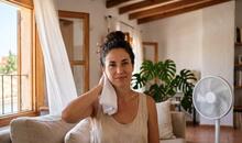 Mujer joven en un salón de casa se refresca el cuello con una toalla blanca junto a un ventilador para aliviar el calor, en representación de la aparición de un sarpullido por calor