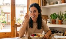 Una mujer sentada en la mesa de una cocina comiendo un bol de yogur natural, alimento rico en postbióticos, junto a frutas y semillas.